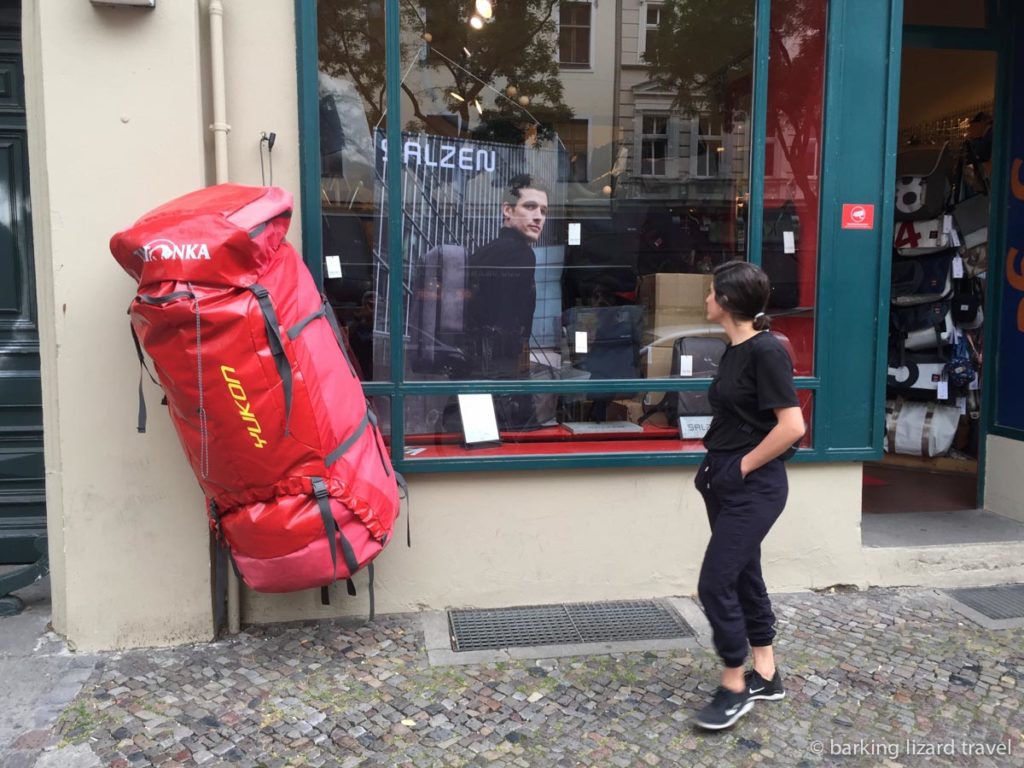 An oversized red backpack hanging up outside a shop. A women in black clothes is walking by